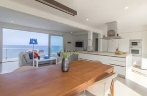Cocina moderna con armarios blancos, una mesa de comedor de madera y vista al mar a través de amplios ventanales.