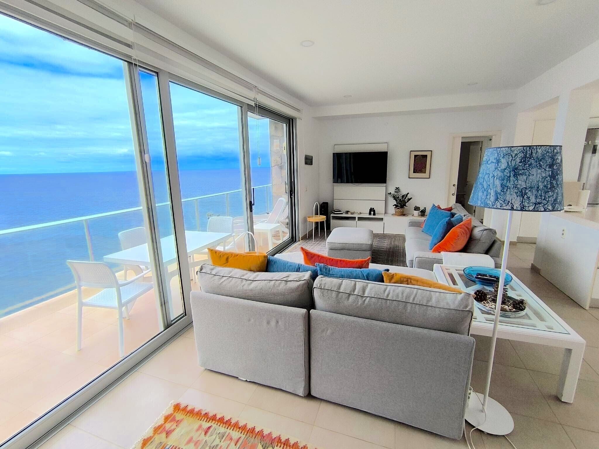 Una sala de estar moderna con un sofá grande, una mesa de centro y un televisor. Hay un balcón con vista al mar.