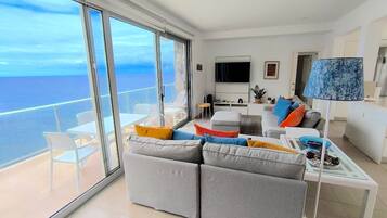Una sala de estar moderna con un sofá grande, una mesa de centro y un televisor. Hay un balcón con vista al mar.