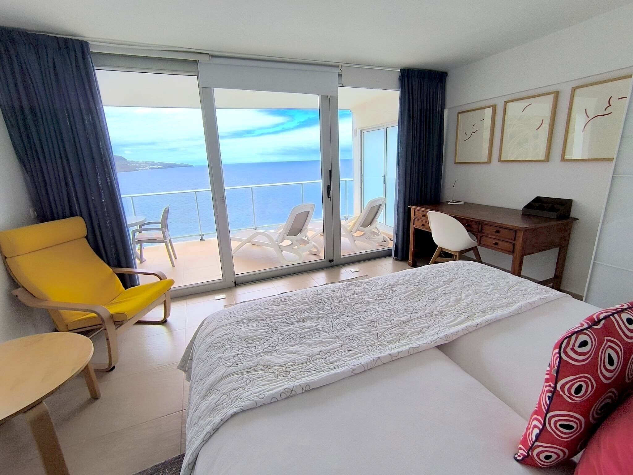 Habitación de hotel con cama, escritorio, silla y vista al mar a través de una puerta corrediza de vidrio.
