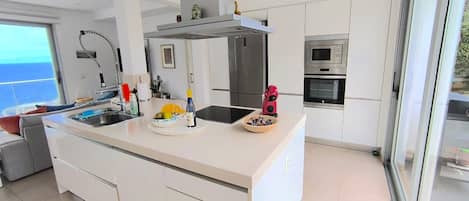 A modern kitchen with a central island, stainless steel appliances, and a view of the sea through a large window.