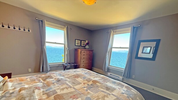 Une chambre à coucher avec un grand lit, deux fenêtres avec des rideaux, une commode et un luminaire fixé au mur.