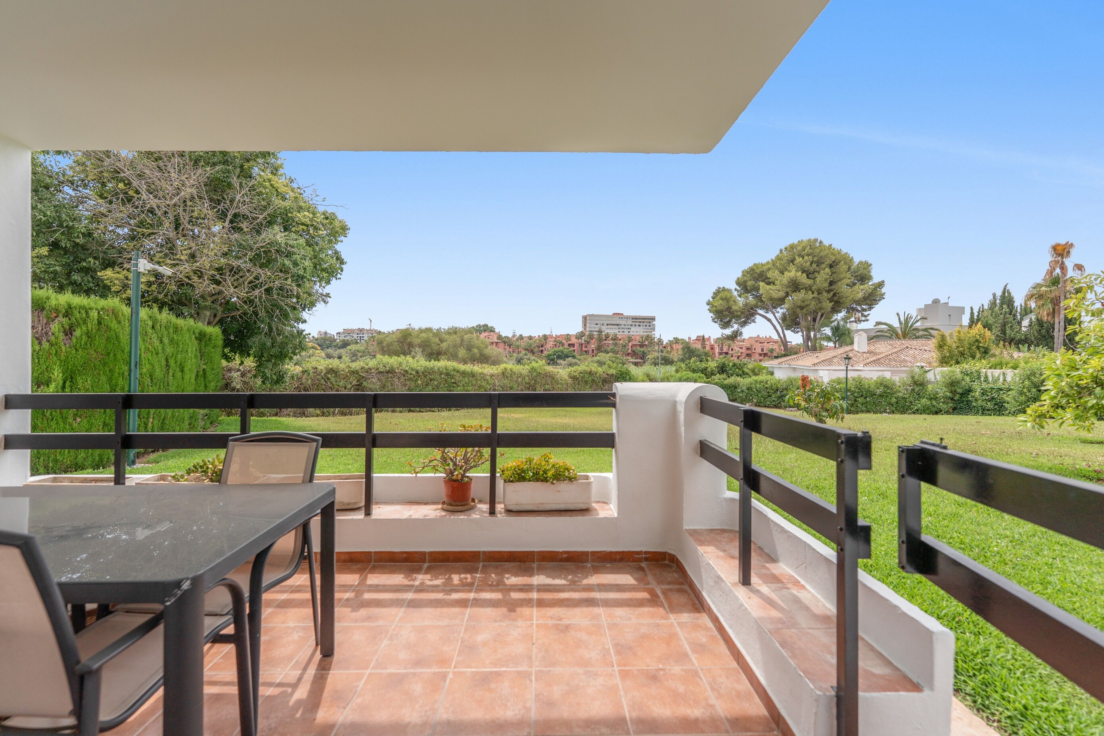 A balcony with a table and chairs, a potted plant, and a view of a green landscape and buildings in the distance.