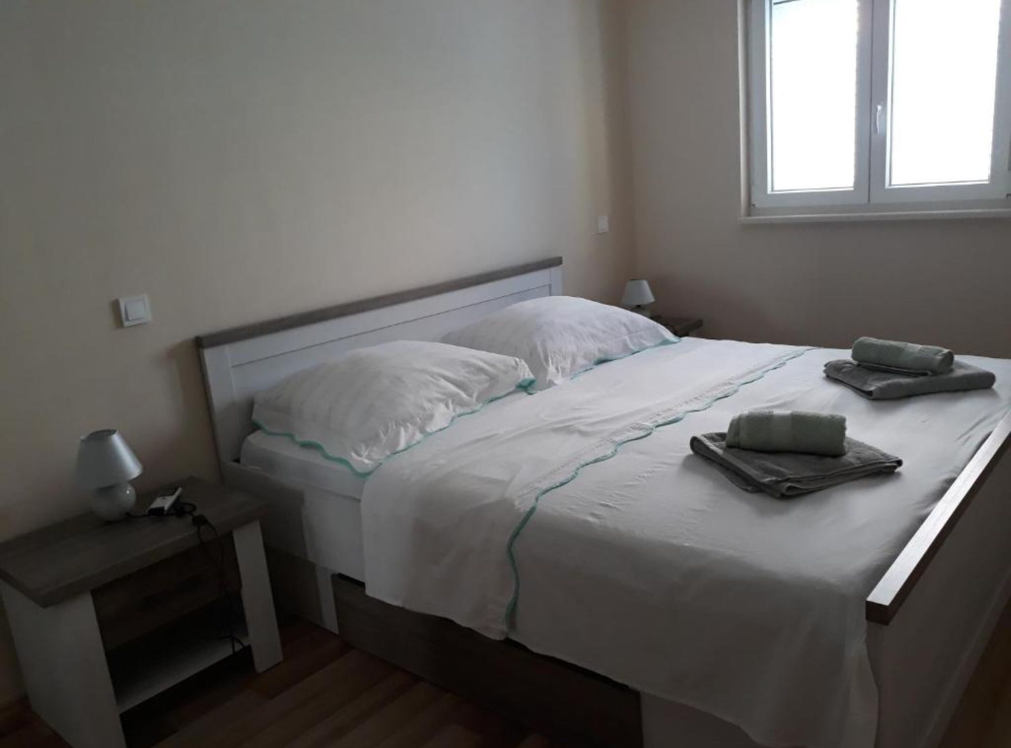 A neatly made bed with pillows and a blanket, a bedside table with a lamp, and a window with daylight.