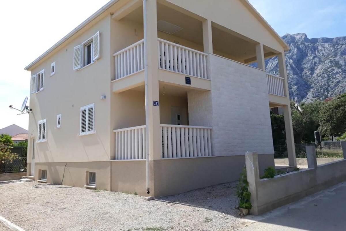 A two-story building with balconies, a gravel driveway, and a mountainous backdrop.
