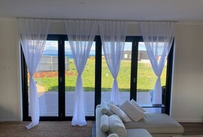 Ein Zimmer mit einer großen Schiebeglaswand, weißen Vorhängen, einer Couch mit Kissen und Blick auf eine Rasenfläche und Berge.
