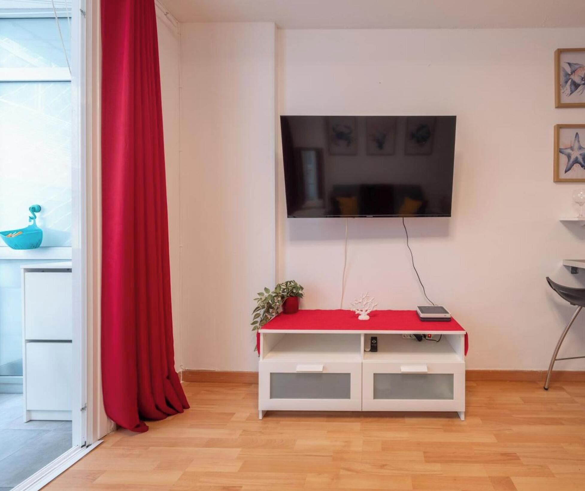 Ein Wohnzimmer mit an der Wand montiertem Fernseher, einem roten Vorhang, einer weißen Unterhaltungsanlage und einem Blumentopf.