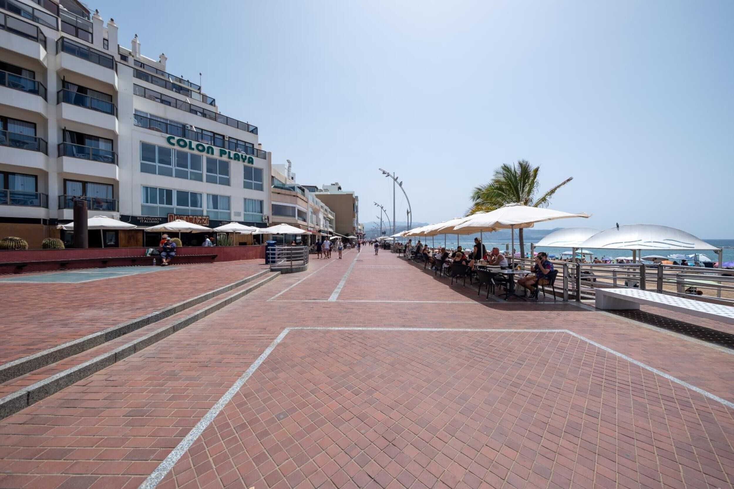 A waterfront promenade with outdoor seating, a restaurant named 'Colon Playa,' and a building with multiple floors and balconies.