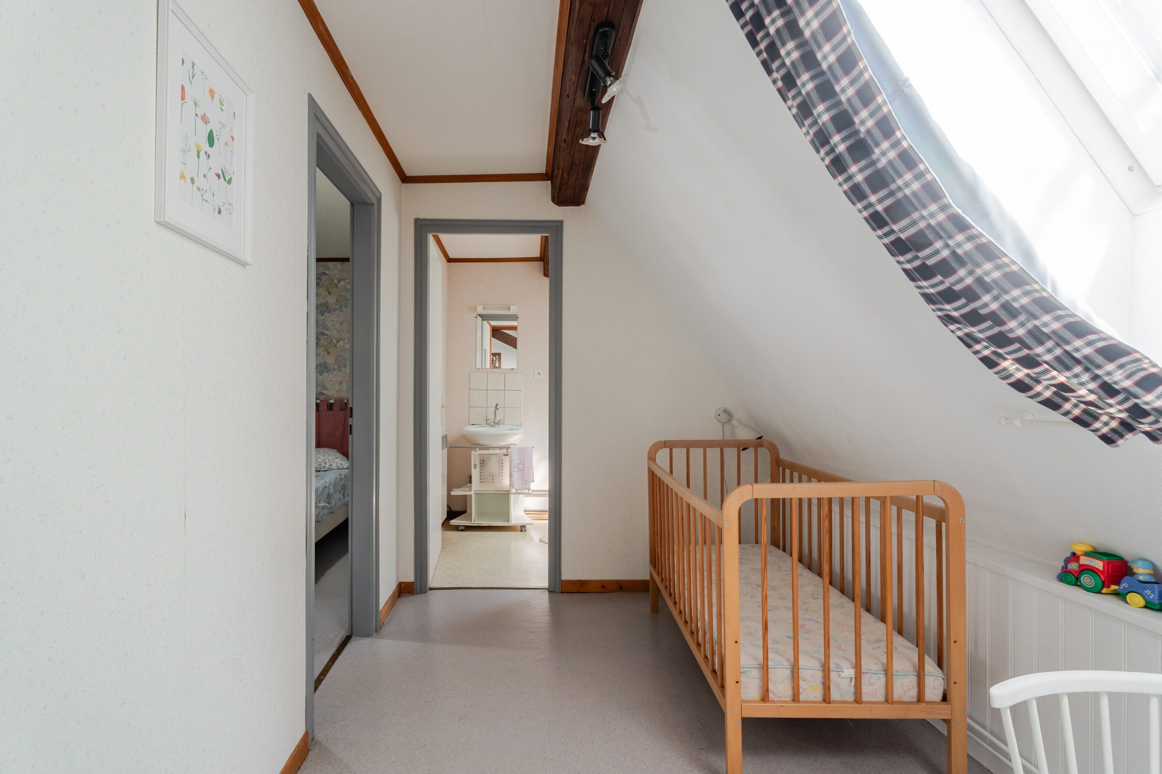 Ein Zimmer mit einem Holckrippe, einem weißen Stuhl, einem Fenster mit Vorhang in Karomuster und einem zur Toilette führenden Gang.
