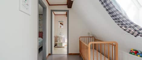 Ein Zimmer mit einem Holckrippe, einem weißen Stuhl, einem Fenster mit Vorhang in Karomuster und einem zur Toilette führenden Gang.