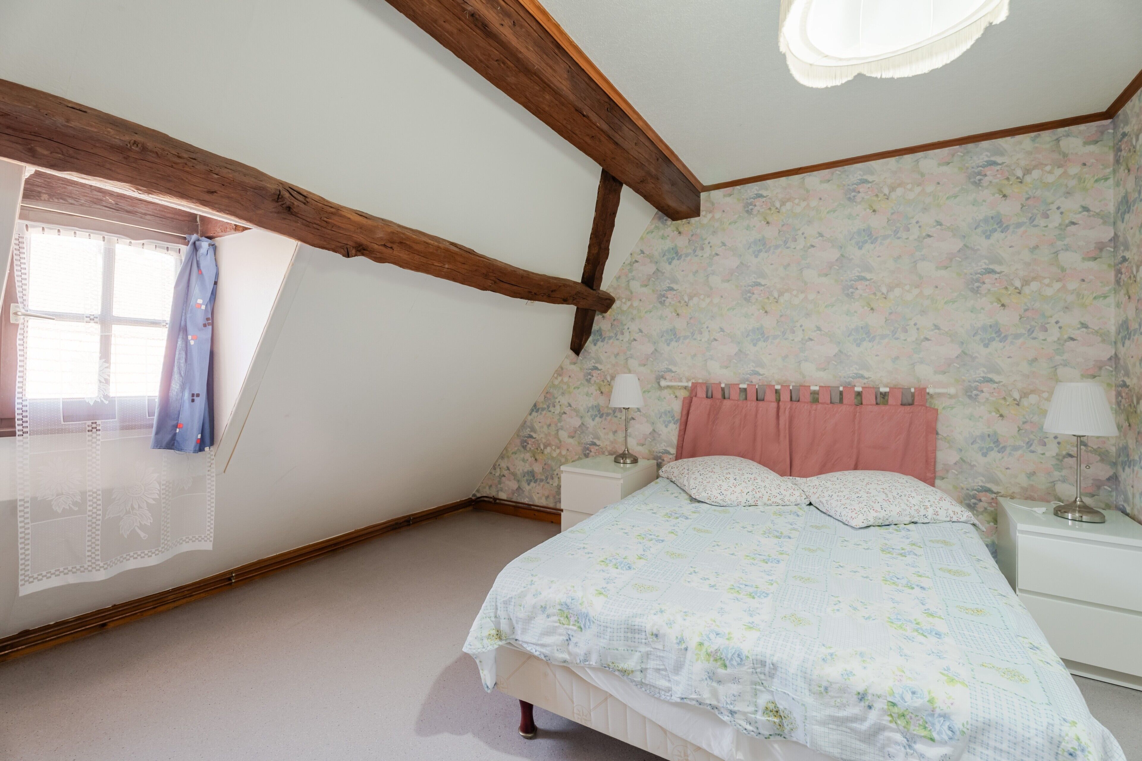 A bedroom with a sloped ceiling, a bed with a floral bedspread, a nightstand with a lamp, and a window with curtains.