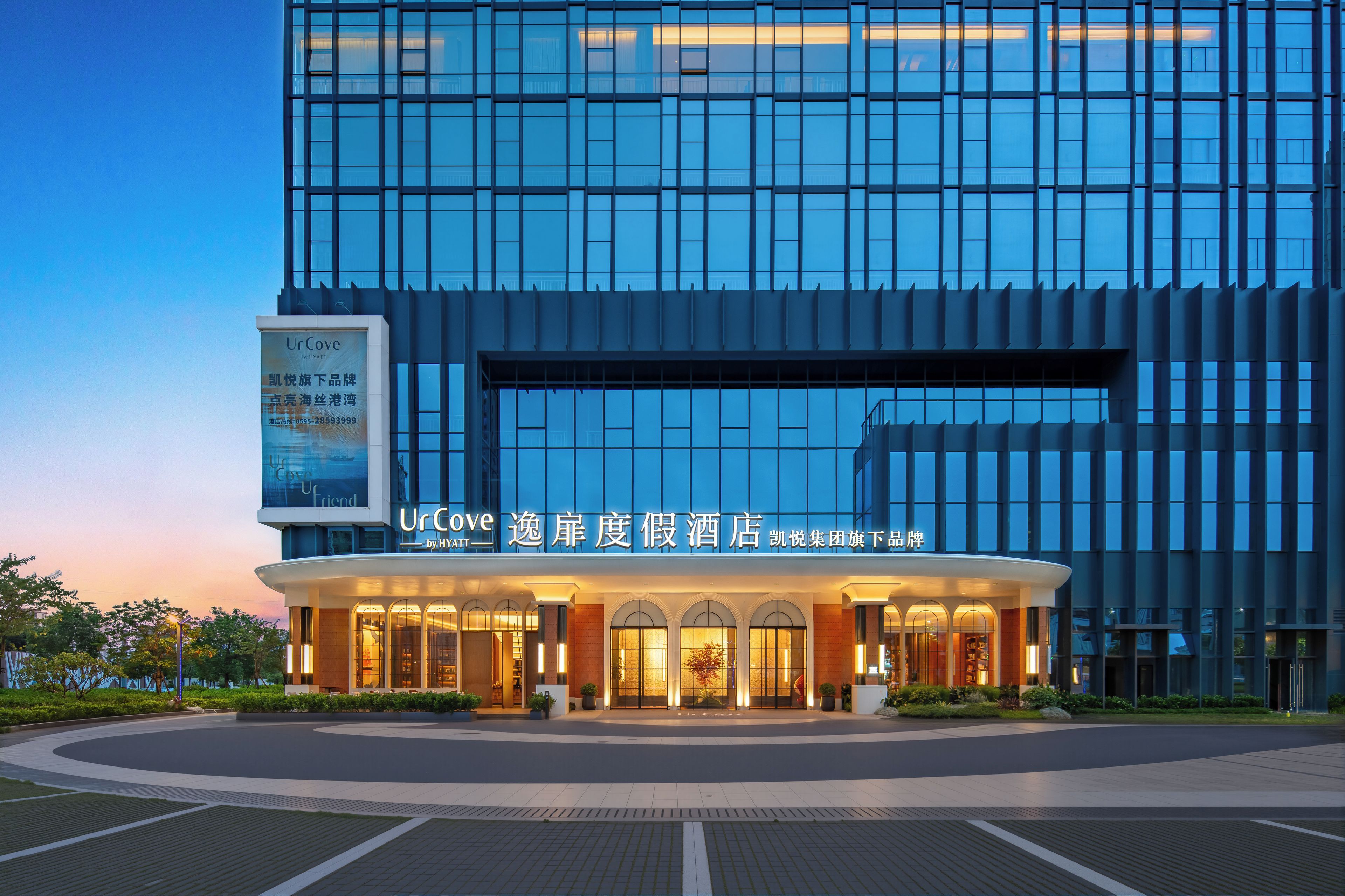 A modern hotel with a glass facade, a well-lit entrance, and a signboard advertising the hotel.