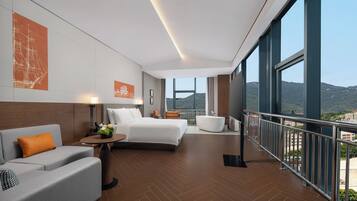 A modern hotel room with a large bed, a sofa, a small table with a vase, and a balcony with a view of the mountains.