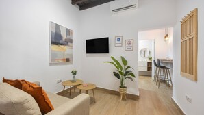Ein Wohnzimmer mit einer Couch, einem Fernseher, einem kleinen Tisch mit einer Pflanze und einem Essbereich mit Stühlen.