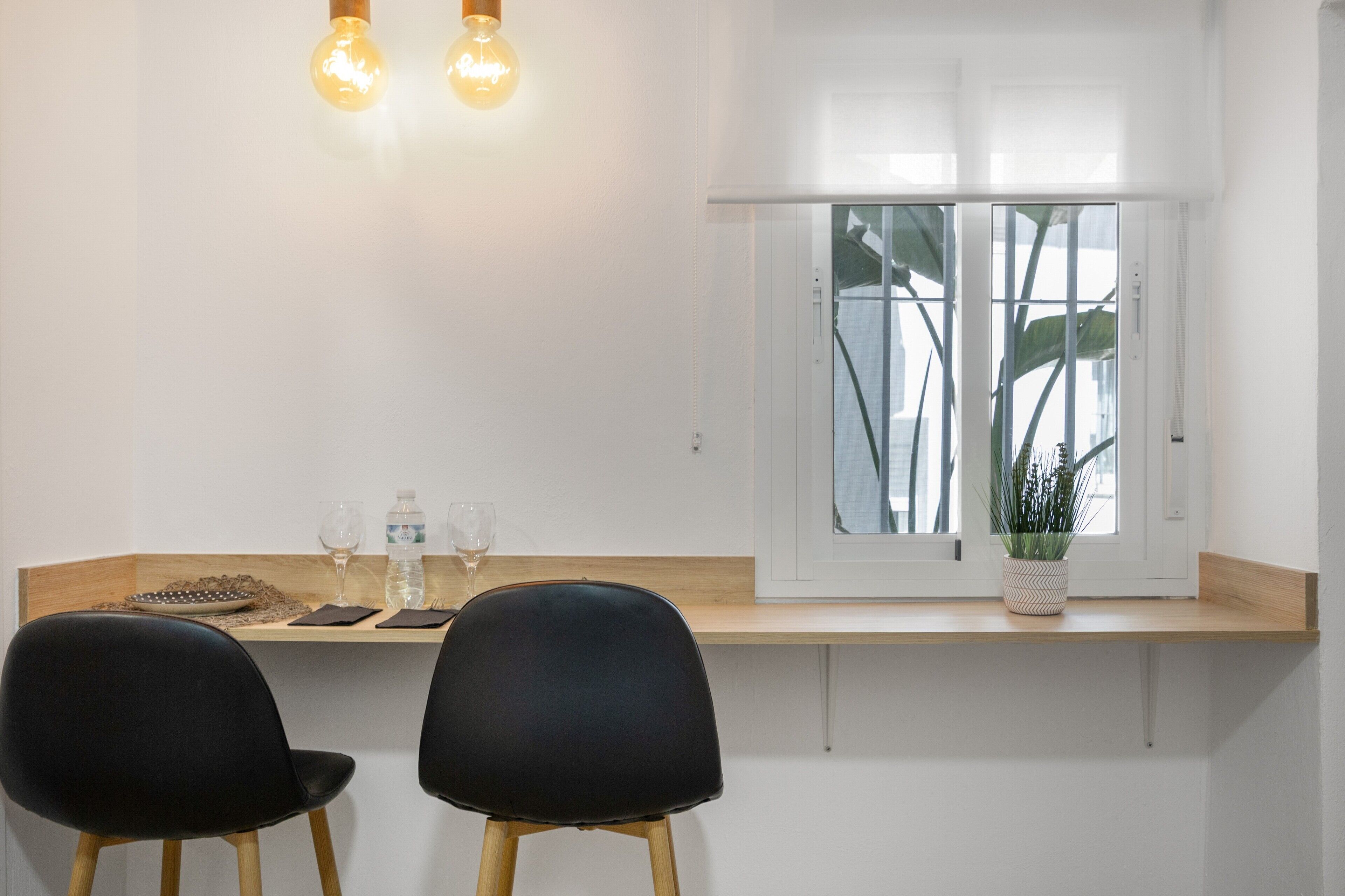 A modern dining area with two black chairs, a wooden table, two wine glasses, a bottle of water, and a potted plant by a window with a view.