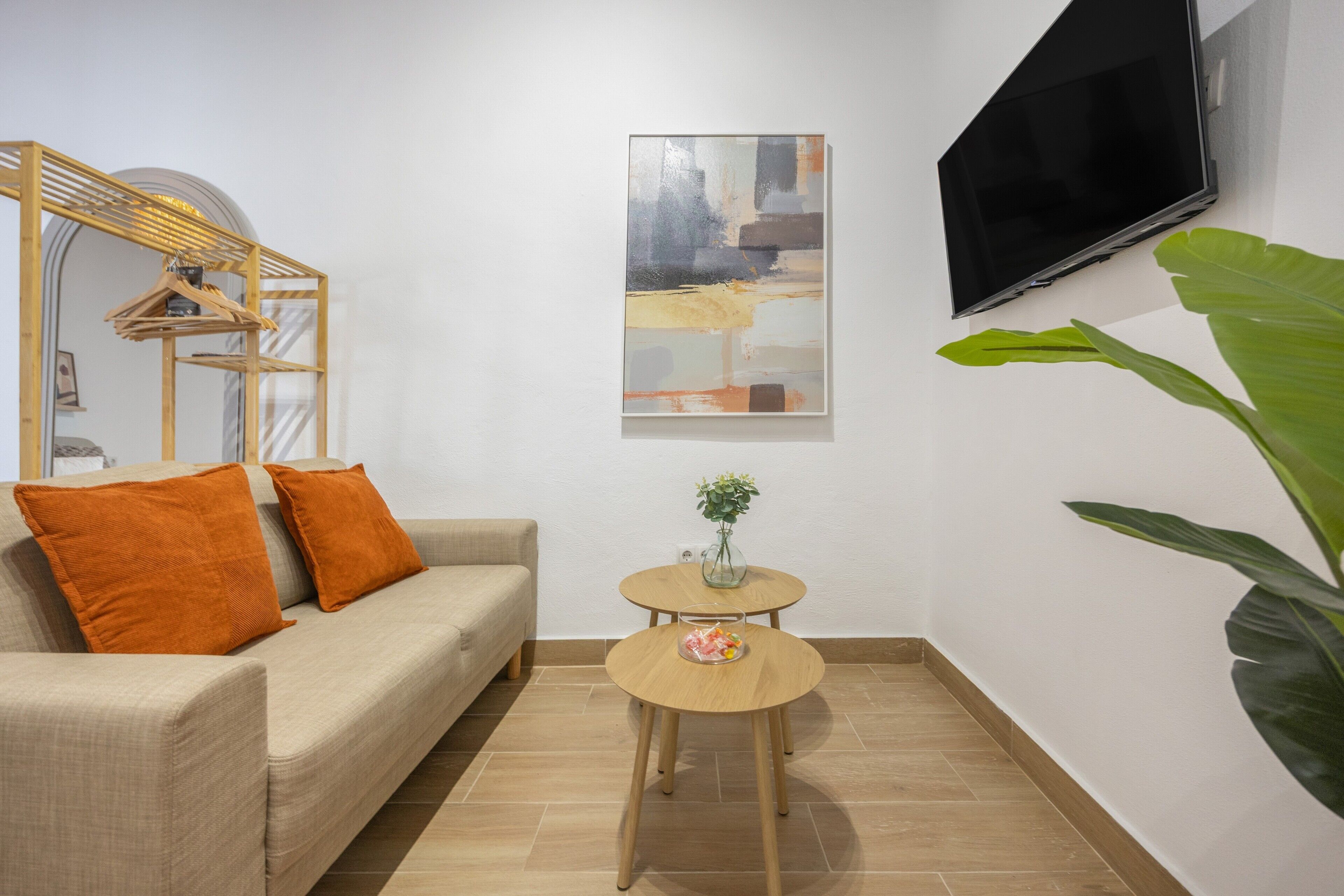 A modern living room with a sofa, a small round table, a flat-screen TV mounted on the wall, and a large green plant.