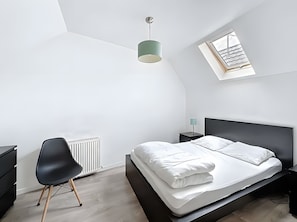 Un dormitorio moderno con una cama, una silla, una mesita de noche, una lámpara, un radiador y un tragaluz.
