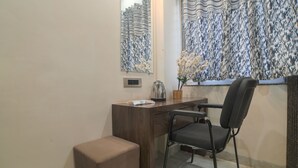 A room with a wooden desk, a chair, a small table with a vase of flowers, a mirror, and patterned curtains. - Shivashish Resort and Marriage Garden (Indore)