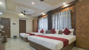 A hotel room with two beds, a desk, a chair, a ceiling fan, and a window with patterned curtains. - Shivashish Resort and Marriage Garden (Indore)