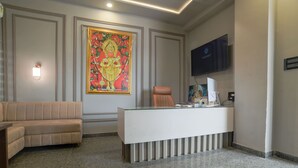 A reception area with a sofa, a reception desk, a television, and a large painting on the wall. - Shivashish Resort and Marriage Garden (Indore)