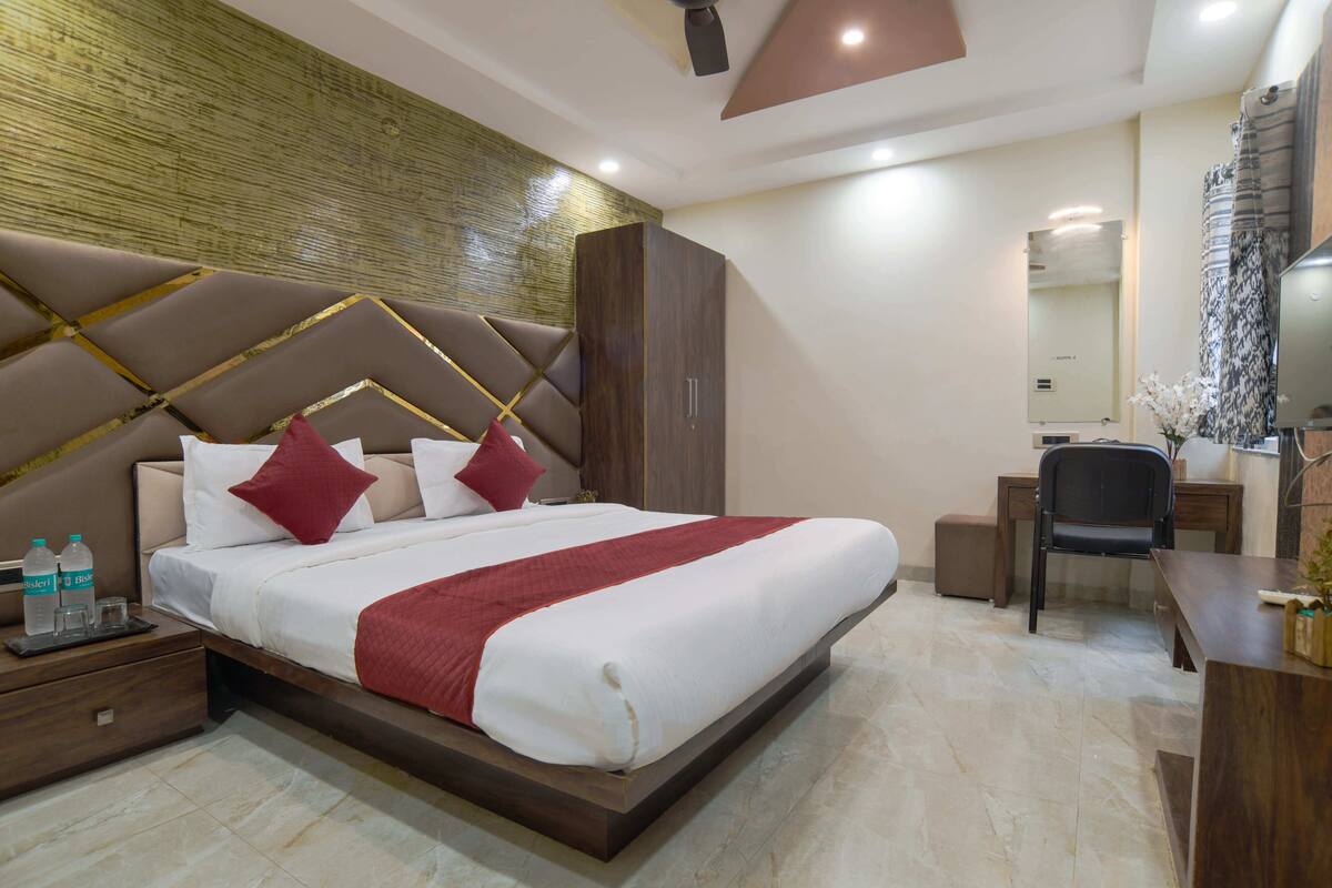 A modern hotel room with a large bed, a wooden wardrobe, a desk with a chair, and a mirror.