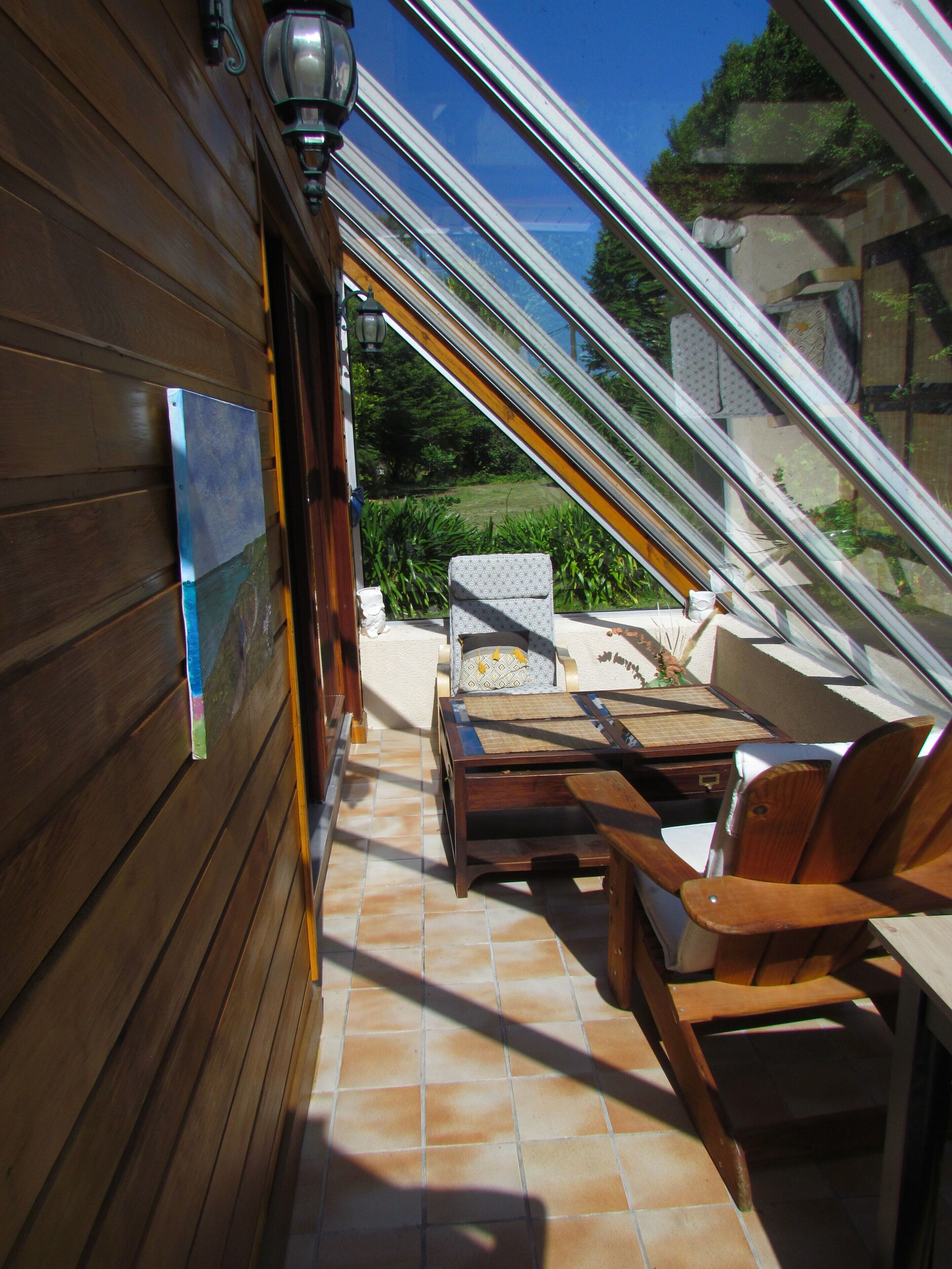 Eine sonnendurchflutete Terrasse mit Holzwänden, einem Tisch, zwei Stühlen und Blick auf Grün hinter einer Glasdachkonstruktion.