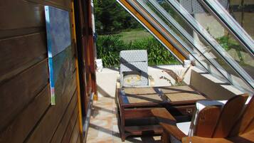 Eine sonnendurchflutete Terrasse mit Holzwänden, einem Tisch, zwei Stühlen und Blick auf Grün hinter einer Glasdachkonstruktion.