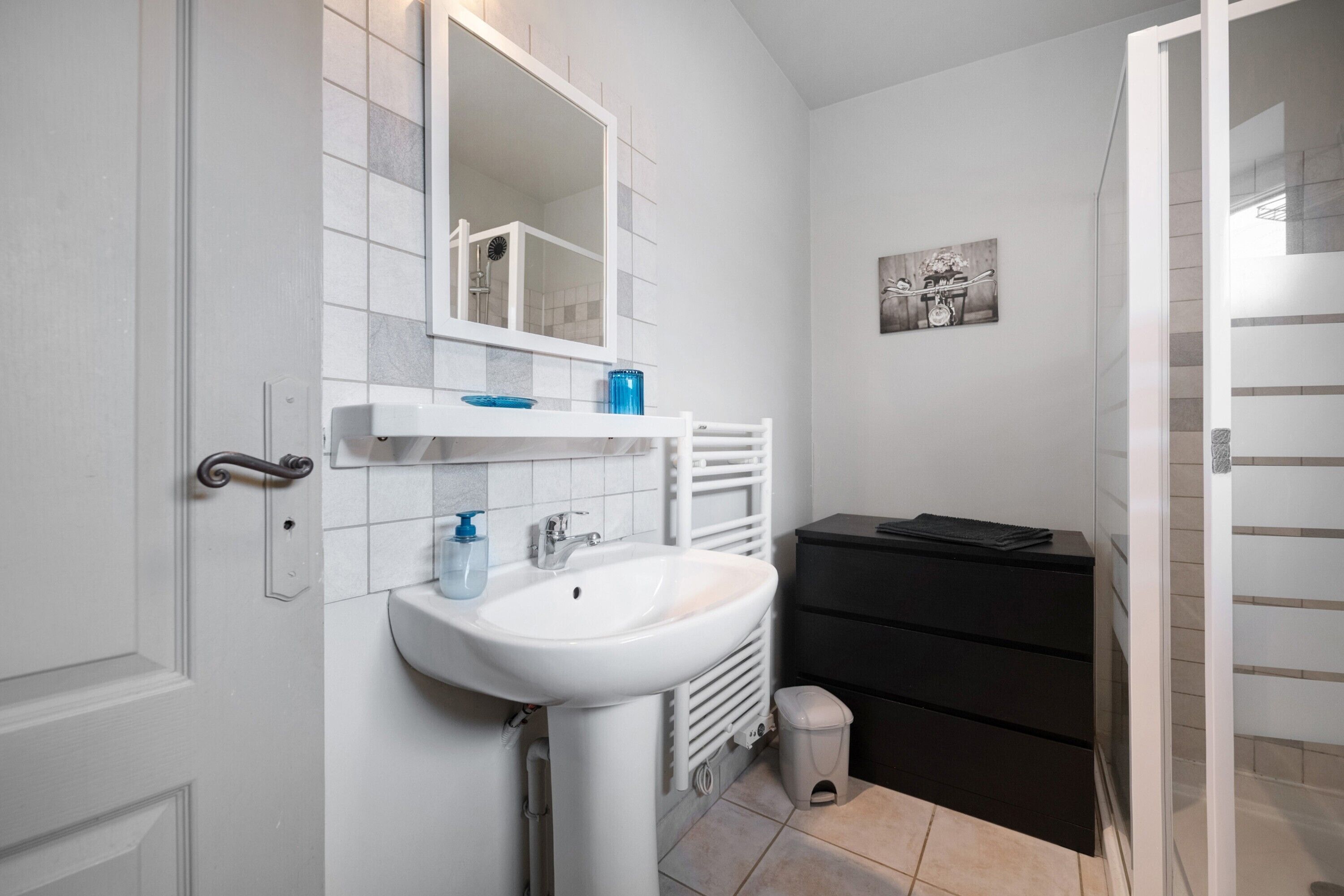 Een badkamer met een witte wasbak, een spiegel, een zwart kastje en een kleine prullenbak.