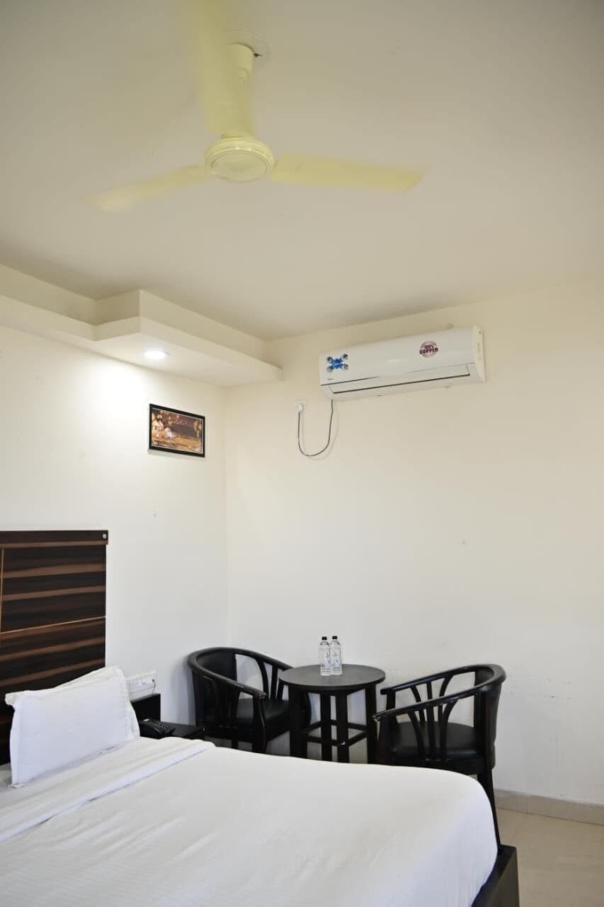 A hotel room with a bed, a ceiling fan, an air conditioning unit, a small table with chairs, and a framed picture on the wall.