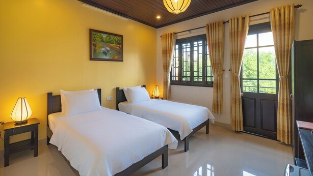 A hotel room with two beds, a painting on the wall, a window with curtains, and a ceiling light. - Old Lane Hoi An Homestay (Hoi An)