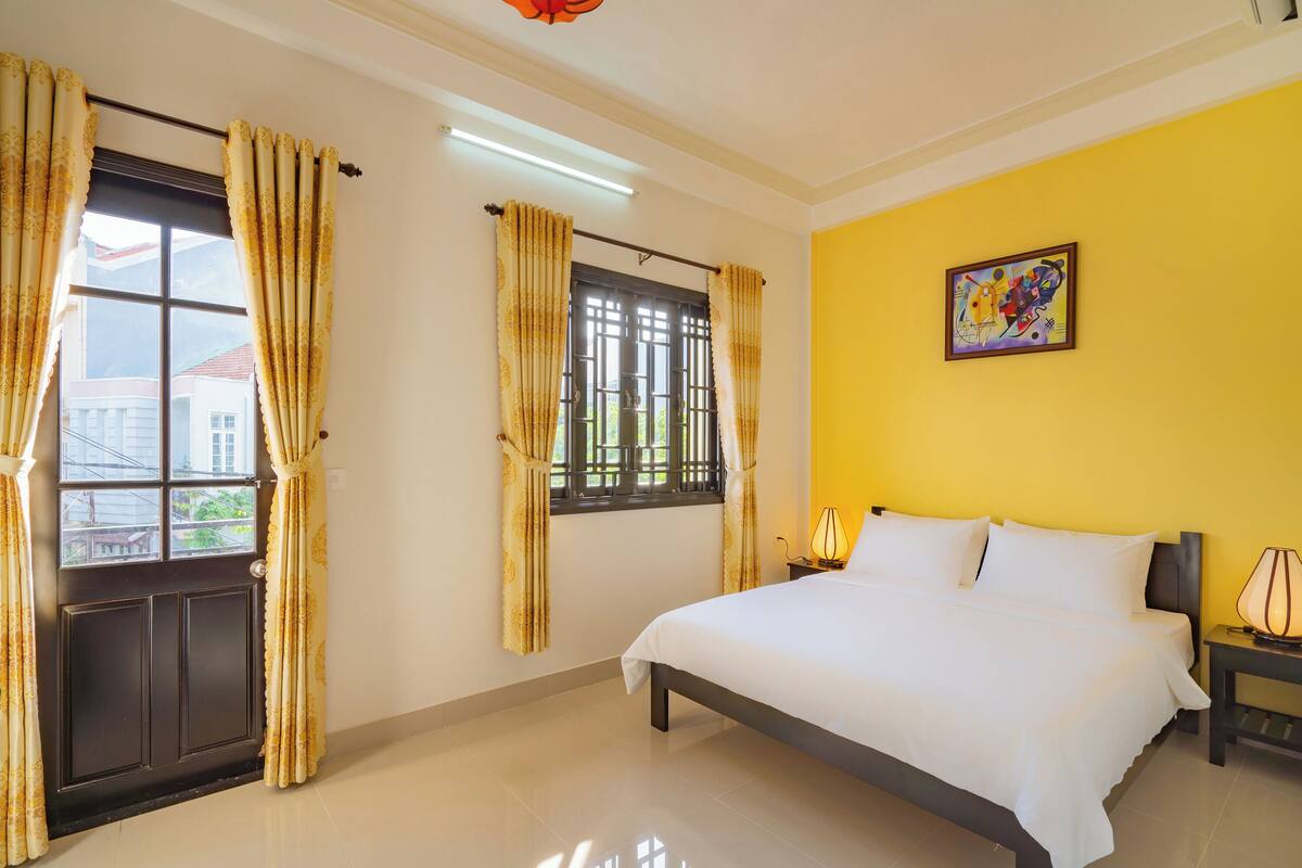 A bedroom with a bed, a nightstand, a lamp, a window with curtains, and a painting on the wall.