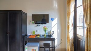 A room with a desk, a television, a cabinet, a window with curtains, and a small table with a vase and a kettle. - Old Lane Hoi An Homestay (Hoi An)