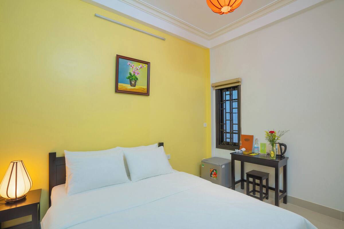 A hotel room with a bed, a desk, a chair, a lamp, a window, and a painting on the wall.