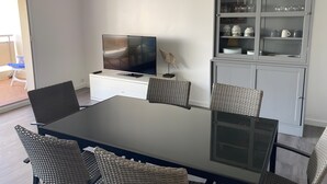Un comedor con una mesa de vidrio, sillas de mimbre, un televisor sobre un mueble blanco y un aparador de vidrio con utensilios de cocina.