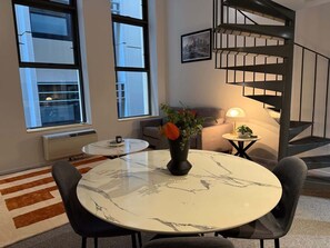 Ein modernes Wohnzimmer mit einem Marmortisch, zwei Stühlen, einer Couch, einem kleinen Beistelltisch mit einer Lampe, einer Vase mit Blumen und einer Treppe.