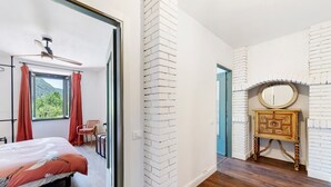 Una habitación con una cama, una ventana con cortinas, una pared de ladrillo, piso de madera y una mesita con un espejo.