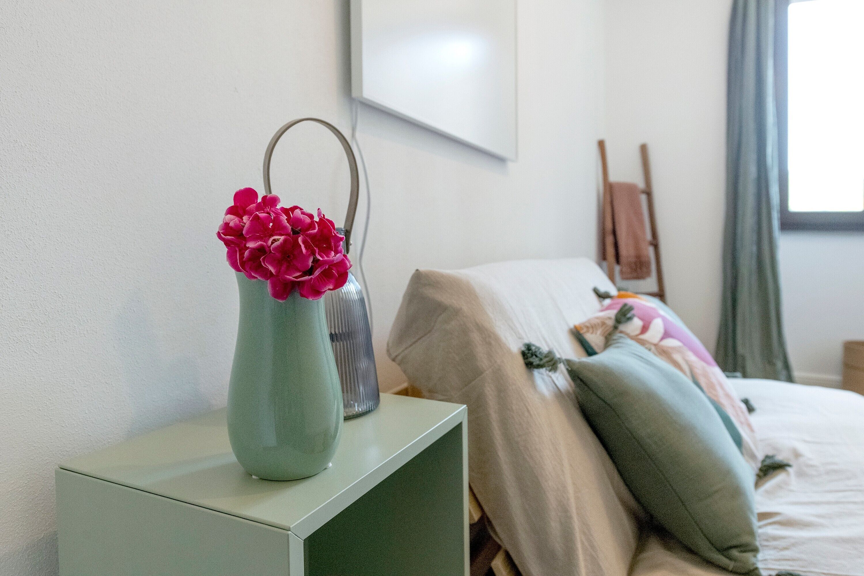 Ein Zimmer mit einem Bett, einem Nachttisch mit Blumen in einer Vase, einem gerahmten Bild an der Wand und einem Fenster mit Vorhängen.