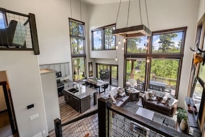 A spacious kitchen with a central island, large windows offering views of greenery, and a dining area with a high ceiling and pendant lighting. - Twilight Pines Cabin (Lead)