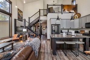 A spacious living area with a large brown leather couch, a staircase, a kitchen with a central island, and a dining area with bar stools. - Twilight Pines Cabin (Lead)