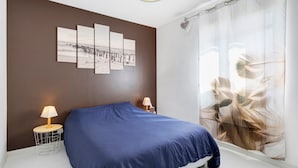 Une chambre à coucher avec un lit, deux tables de chevet avec des lampes, un mur orné d’un grand tableau abstrait et une fenêtre avec des rideaux légers.