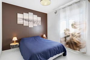 A bedroom with a bed, two bedside tables with lamps, a wall with a large abstract painting, and a window with sheer curtains.