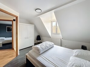 Une chambre avec un plafond en pente, un lit avec une literie blanche, une chaise noire et une armoire.