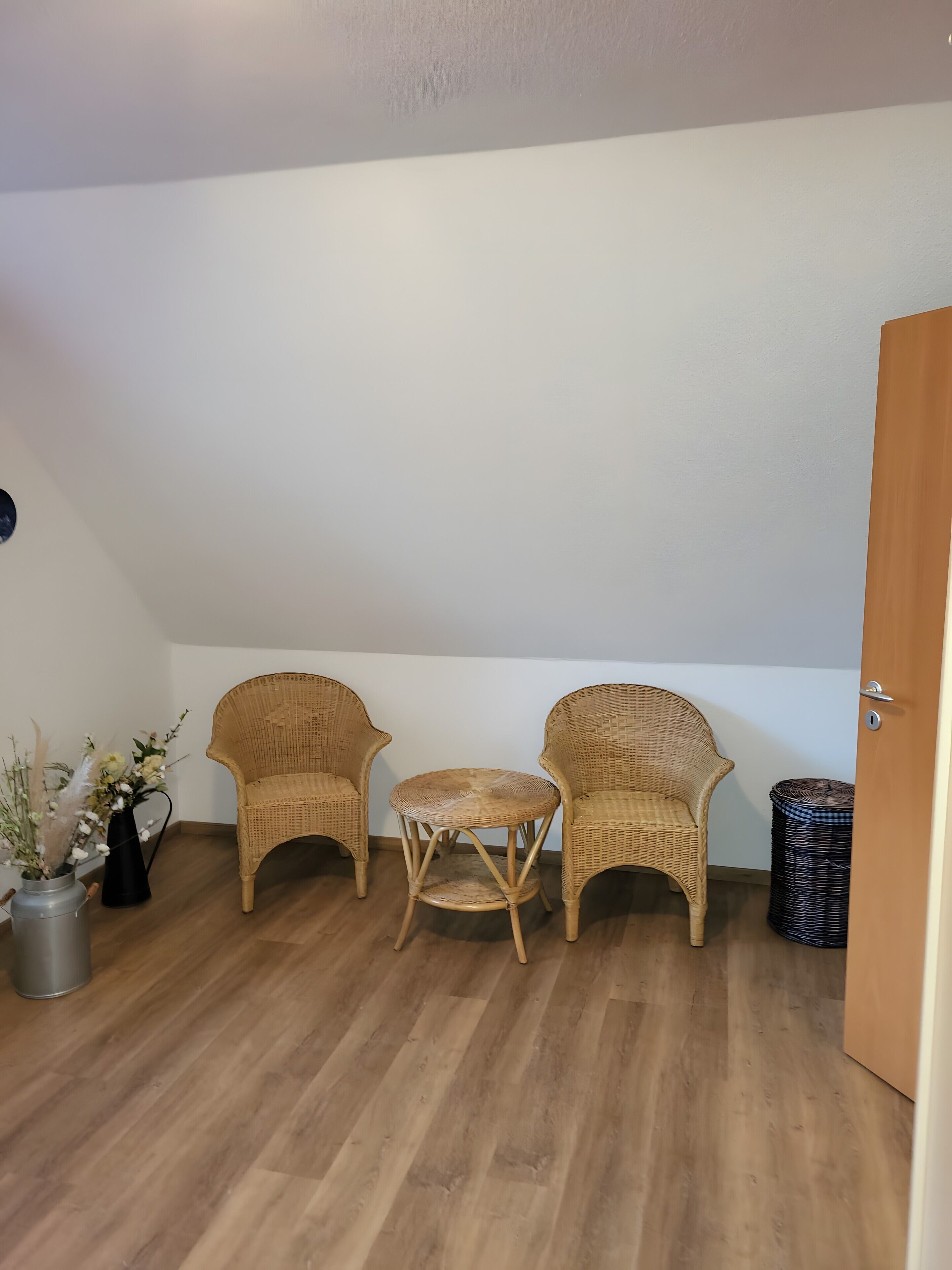 Ein Zimmer mit zwei Korbstühlen, einem kleinen runden Tisch, einem schwarzen Korbkorb und einem silbernen Mülleimer.