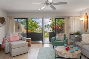 Ein modernes Wohnzimmer mit Deckenventilator, zwei Sofas, einem Couchtisch und Blick auf Palmen durch große Glasfenster.
