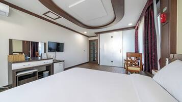 Quarto de hotel com cama, escrivaninha com computador, televisão, guarda-roupa e uma cadeira.