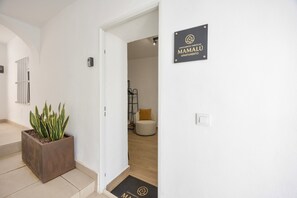 Un pasillo con una planta en maceta, un letrero que indica 'MAMALDU APARTMENT' y un felpudo con el mismo logotipo.