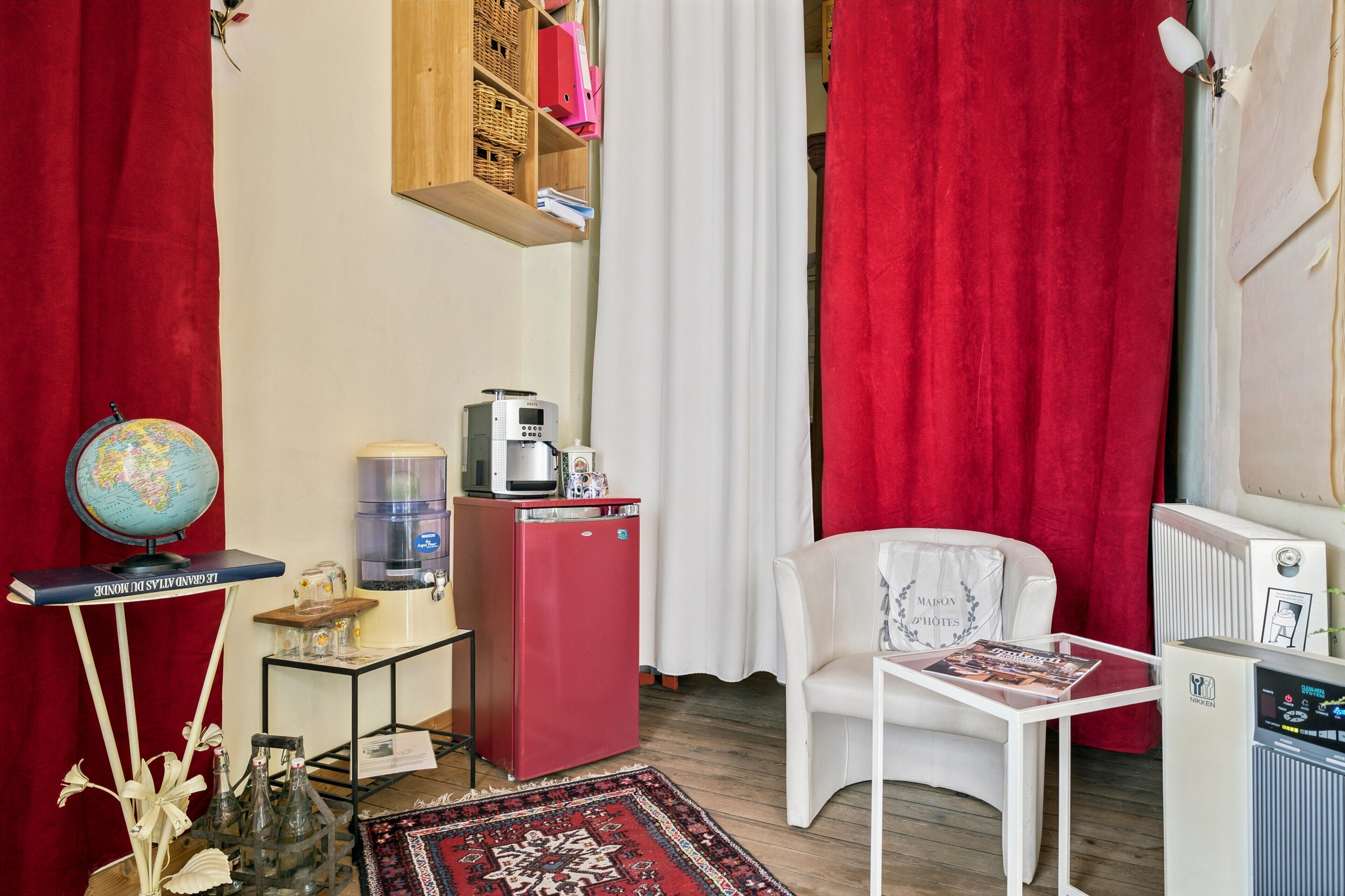 A compact room with a red refrigerator, a white chair, a small table with a magazine, a globe, and a water dispenser.