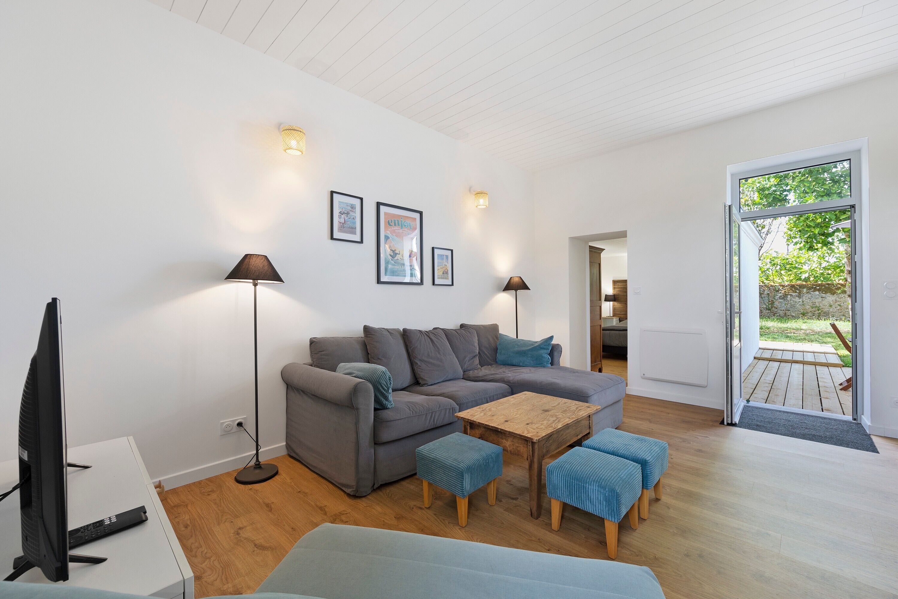 Ein modernes Wohnzimmer mit einem grauen Sofa, einem Holztisch und blauen Hocker. Zwei Stehlampen, ein Fernseher und eine Tür zu einem Außenbereich sind ebenfalls vorhanden.