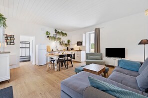 A modern living room with a grey sofa, a TV, and a dining area with a kitchen in the background.