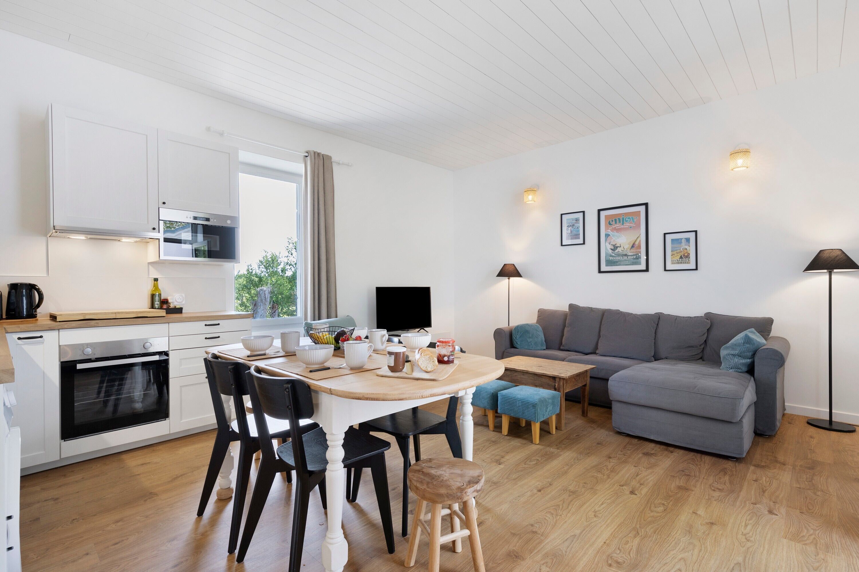 Ett modernt kök med vita skåp, ett matbord med stolar, en grå soffa och en tv.